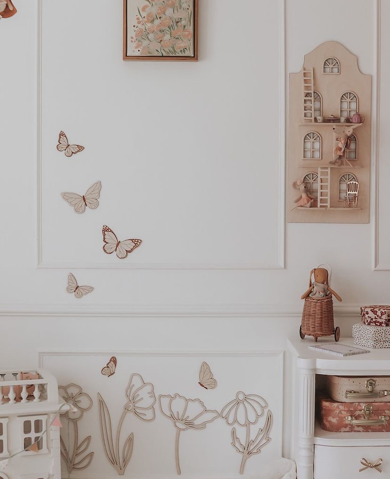 May include: A light-toned nursery scene with wooden butterfly and flower wall decorations. A wooden dollhouse and a decorative house-shaped shelf with miniature figures are also visible. A doll in a wicker cart and stacked suitcases complete the decor.