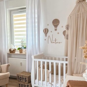 Puede incluir: Una cuna blanca con una s&aacute;bana estampada en blanco y gris y un parachoques beige. La cuna est&aacute; en una habitaci&oacute;n de beb&eacute; con cortinas blancas, una cesta de mimbre blanca con la palabra "TOYS" en ella y un dosel beige con pompones colgando. La pared est&aacute; decorada con globos aerost&aacute;ticos de madera y el nombre "Natan" escrito en cursiva.