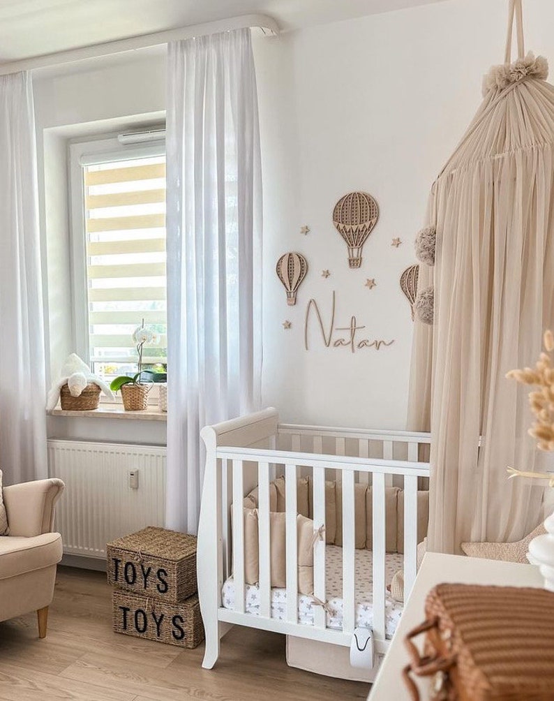 Puede incluir: Una cuna blanca con una s&aacute;bana estampada en blanco y gris y un parachoques beige. La cuna est&aacute; en una habitaci&oacute;n de beb&eacute; con cortinas blancas, una cesta de mimbre blanca con la palabra "TOYS" en ella y un dosel beige con pompones colgando. La pared est&aacute; decorada con globos aerost&aacute;ticos de madera y el nombre "Natan" escrito en cursiva.