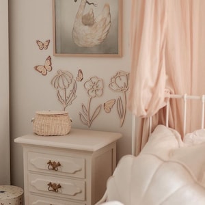 May include: A bedroom scene with a white nightstand with three drawers, a woven basket, and wooden flower and butterfly wall decorations. A framed swan print hangs above the nightstand. A bed with a pink canopy is in the background.