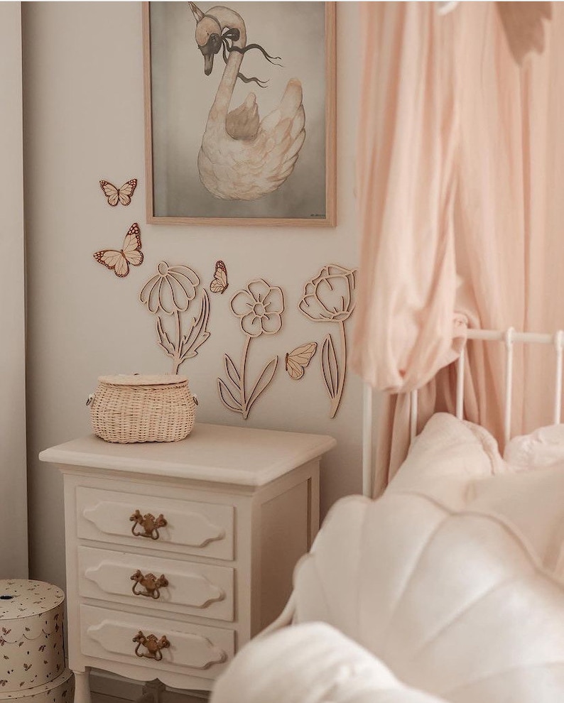 May include: A bedroom scene with a white nightstand with three drawers, a woven basket, and wooden flower and butterfly wall decorations. A framed swan print hangs above the nightstand. A bed with a pink canopy is in the background.