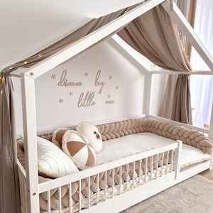 K&ouml;nnte beinhalten: Wei&szlig;es Holzbettgestell mit Baldachin und einem Baldachin aus beigefarbenem Stoff. Das Bett hat einen geflochtenen Sto&szlig;f&auml;nger in Beige und eine wei&szlig;e Matratze. Die Wand hinter dem Bett hat die Worte "dream big little one" in goldenen Buchstaben geschrieben.