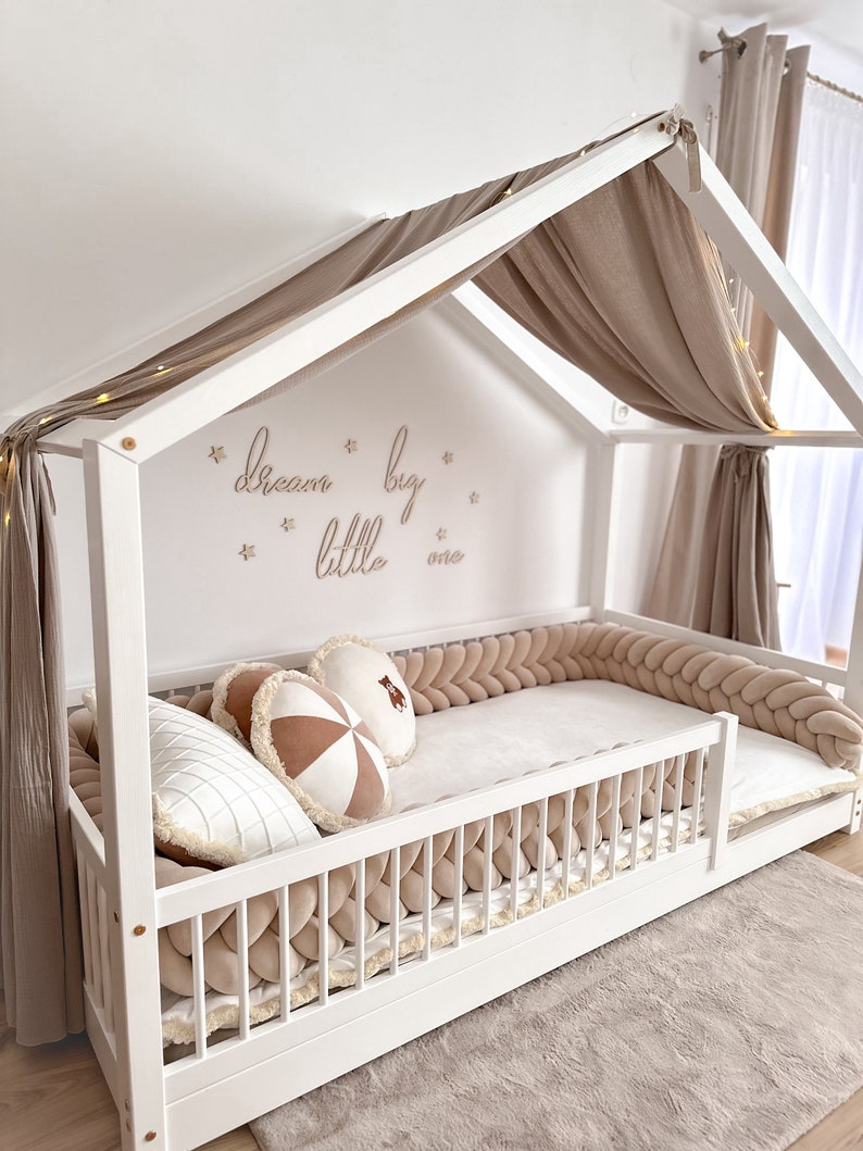 K&ouml;nnte beinhalten: Wei&szlig;es Holzbettgestell mit Baldachin und einem Baldachin aus beigefarbenem Stoff. Das Bett hat einen geflochtenen Sto&szlig;f&auml;nger in Beige und eine wei&szlig;e Matratze. Die Wand hinter dem Bett hat die Worte "dream big little one" in goldenen Buchstaben geschrieben.