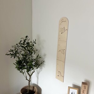 K&ouml;nnte beinhalten: Eine h&ouml;lzerne Wachstumstabelle mit ge&auml;tzten Vogelmotiven und Messmarkierungen. Der Name "Serra" steht oben. Die Tabelle ist an einer wei&szlig;en Wand befestigt, mit einem kleinen Baum in einem geflochtenen Korb und Holzbl&ouml;cken in der N&auml;he.