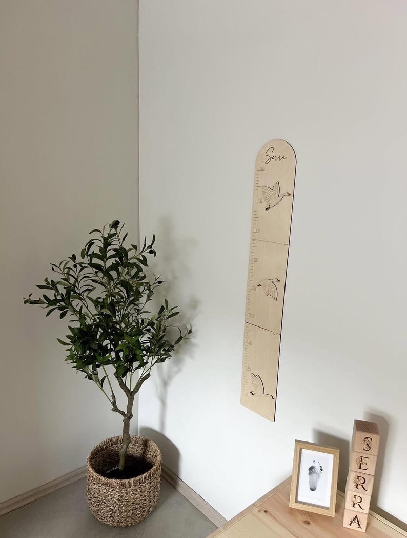 K&ouml;nnte beinhalten: Eine h&ouml;lzerne Wachstumstabelle mit ge&auml;tzten Vogelmotiven und Messmarkierungen. Der Name "Serra" steht oben. Die Tabelle ist an einer wei&szlig;en Wand befestigt, mit einem kleinen Baum in einem geflochtenen Korb und Holzbl&ouml;cken in der N&auml;he.
