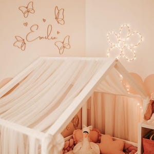 May include: A child's bedroom with a white canopy bed, adorned with a sheer, light-colored fabric. The walls are a soft peach, decorated with wooden butterfly cutouts and the name "Emilia". A flower-shaped light fixture adds a warm glow.