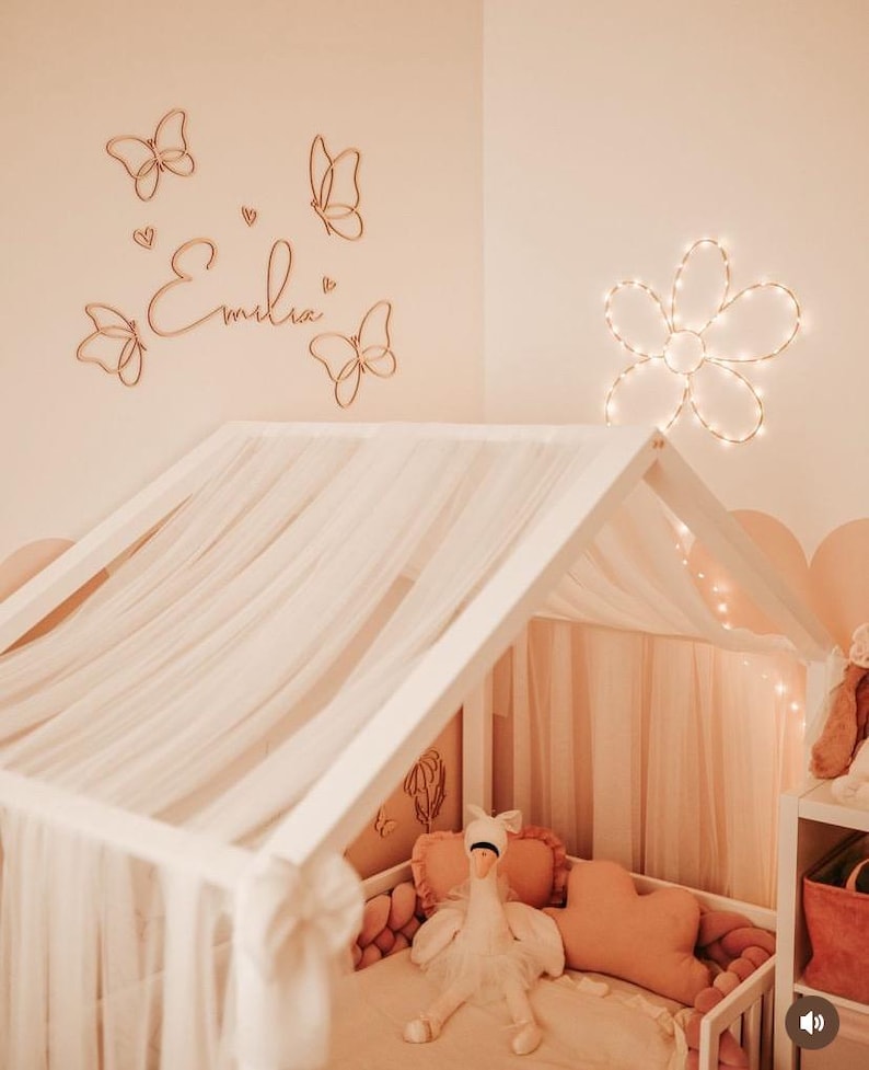 May include: A child's bedroom with a white canopy bed, adorned with a sheer, light-colored fabric. The walls are a soft peach, decorated with wooden butterfly cutouts and the name "Emilia". A flower-shaped light fixture adds a warm glow.