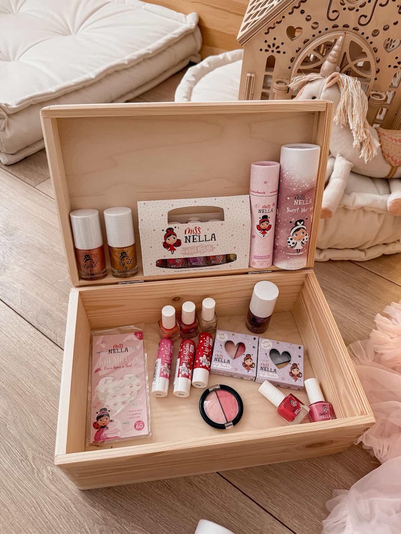 Op de afbeelding: Een open houten kist gevuld met kindermake-upproducten. De kist bevat nagellak, lipgloss, oogschaduw en andere schoonheidsproducten. De producten zijn voorzien van de tekst "miss NELLA" en tonen een cartoonmeisje.