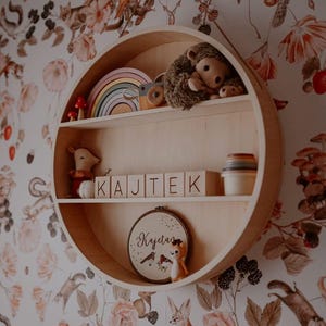 May include: A circular wooden shelf with two shelves, featuring a rainbow stacking toy, a stuffed hedgehog, a wooden toy, and a small stuffed animal. The shelf is decorated with wooden blocks spelling "KAJTEK" and a circular embroidery hoop with the name "Kajetan" in cursive.