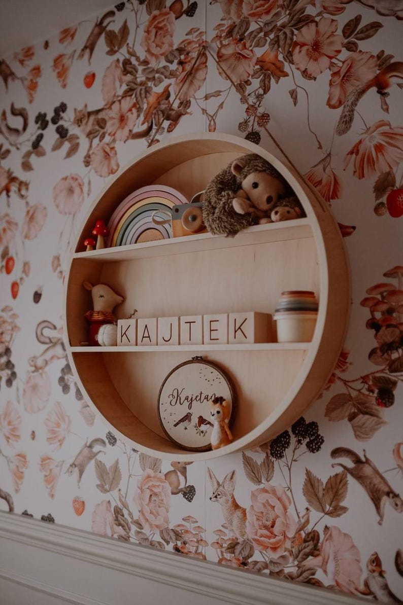 May include: A circular wooden shelf with two shelves, featuring a rainbow stacking toy, a stuffed hedgehog, a wooden toy, and a small stuffed animal. The shelf is decorated with wooden blocks spelling "KAJTEK" and a circular embroidery hoop with the name "Kajetan" in cursive.