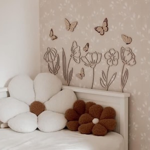 May include: A bedroom scene featuring a white bed with a large, daisy-shaped pillow and a smaller, brown flower pillow. The wall is decorated with wooden butterfly and flower cutouts, creating a whimsical, nature-inspired theme.