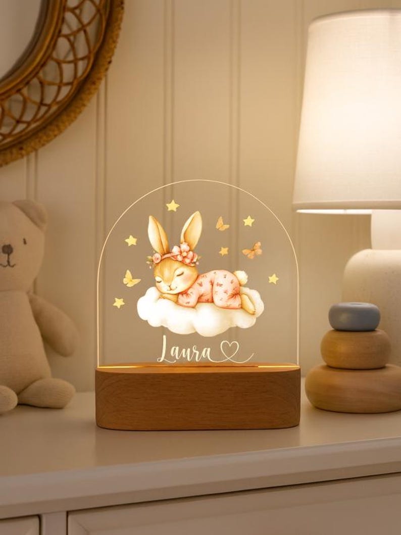 May include: A decorative night light featuring a sleeping bunny on a cloud, with the name "Laura" and a heart. The bunny wears a floral crown and is surrounded by stars and butterflies. The base is made of wood.