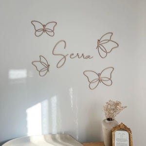May include: Wall decor featuring the name "Serra" in cursive script, accompanied by four delicate butterfly outlines. The butterflies and name are in a light brown color, creating a soft, neutral aesthetic. The arrangement is on a white wall.