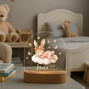 May include: A decorative night light featuring a sleeping bunny on a cloud, with the name "Laura" and a heart. The design is on a clear acrylic arch, set on a wooden base. Butterflies and stars surround the bunny.