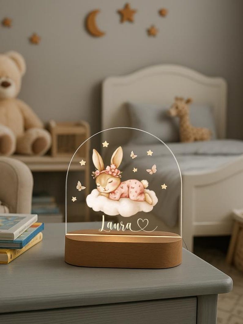 May include: A decorative night light featuring a sleeping bunny on a cloud, with the name "Laura" and a heart. The design is on a clear acrylic arch, set on a wooden base. Butterflies and stars surround the bunny.