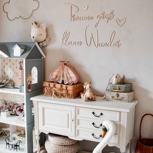 May include: A white dresser with three drawers and black handles. A white swan plush toy sits on a white cushion in front of the dresser. A wicker basket sits on the floor next to the dresser. The wall behind the dresser has a wooden sign that reads "Traume gro&szlig; kleines Wunder".