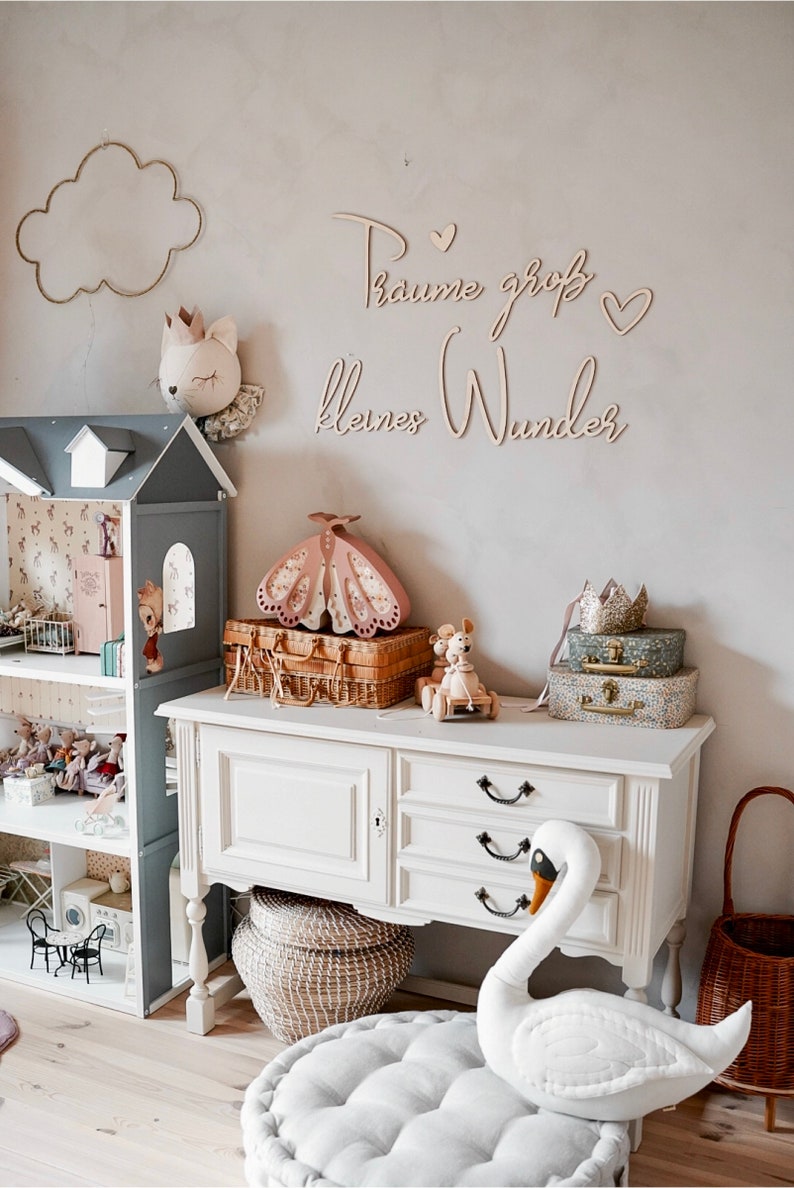 May include: A white dresser with three drawers and black handles. A white swan plush toy sits on a white cushion in front of the dresser. A wicker basket sits on the floor next to the dresser. The wall behind the dresser has a wooden sign that reads "Traume gro&szlig; kleines Wunder".