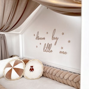 K&ouml;nnte beinhalten: Ein wei&szlig;es Babybett mit einem geflochtenen Sto&szlig;f&auml;nger in Braun und Wei&szlig;. Das Bett hat einen Baldachin aus beigefarbenem Stoff mit wei&szlig;en Lichterketten. Die Wand hinter dem Bett hat ein Holzschild mit der Aufschrift "dream big little one" mit goldenen Sternen.