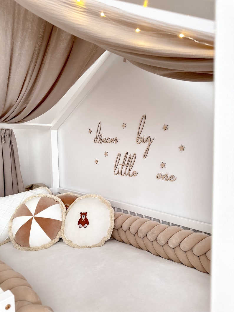 K&ouml;nnte beinhalten: Ein wei&szlig;es Babybett mit einem geflochtenen Sto&szlig;f&auml;nger in Braun und Wei&szlig;. Das Bett hat einen Baldachin aus beigefarbenem Stoff mit wei&szlig;en Lichterketten. Die Wand hinter dem Bett hat ein Holzschild mit der Aufschrift "dream big little one" mit goldenen Sternen.
