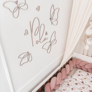 May include: Wooden butterfly wall art and the name "Pola" in cursive script are mounted on a white wall. A crib with a pink braided bumper and floral bedding is in the foreground. A white canopy hangs above the crib.