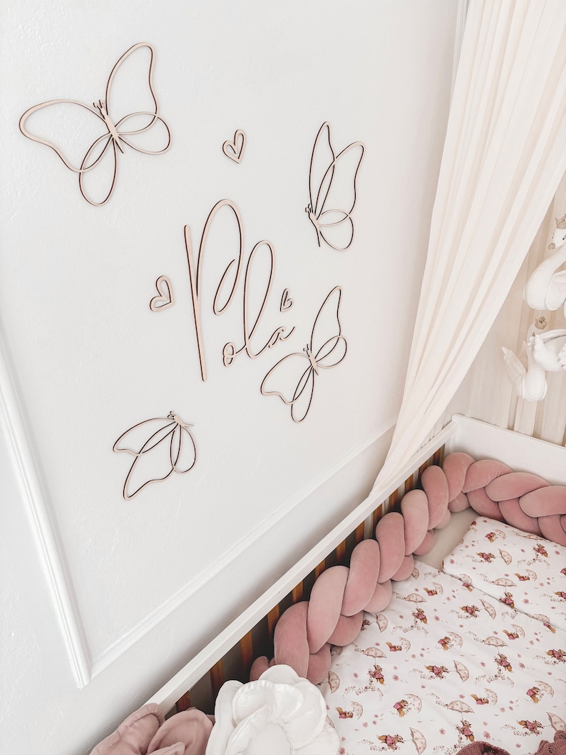 May include: Wooden butterfly wall art and the name "Pola" in cursive script are mounted on a white wall. A crib with a pink braided bumper and floral bedding is in the foreground. A white canopy hangs above the crib.