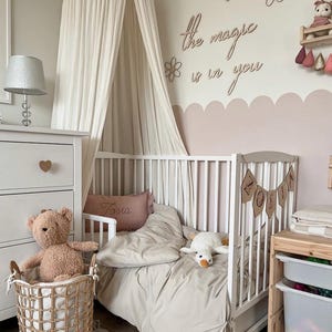 Pu&ograve; includere: Un lettino bianco con una zanzariera bianca e un set di biancheria da letto beige e bianco. Il lettino ha uno stendardo con il nome in legno che dice "ZOSIA". La parete dietro il lettino &egrave; dipinta di rosa chiaro e ha un cartello in legno che dice "La magia &egrave; in te".