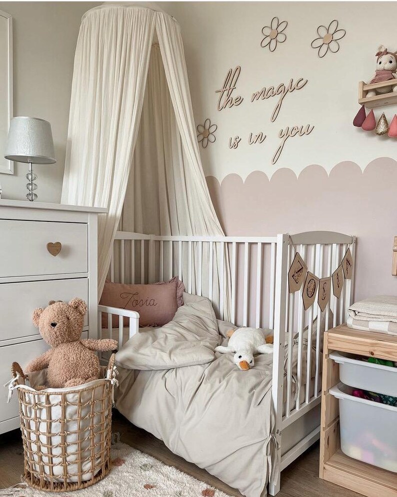 Pu&ograve; includere: Un lettino bianco con una zanzariera bianca e un set di biancheria da letto beige e bianco. Il lettino ha uno stendardo con il nome in legno che dice "ZOSIA". La parete dietro il lettino &egrave; dipinta di rosa chiaro e ha un cartello in legno che dice "La magia &egrave; in te".