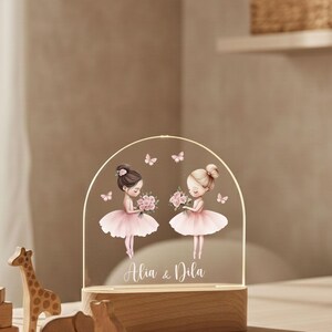 May include: A decorative night light featuring two cartoon ballerinas in pink tutus, holding bouquets, and surrounded by butterflies. The names "Alia & Dila" are written below the ballerinas. The light sits on a wooden base.