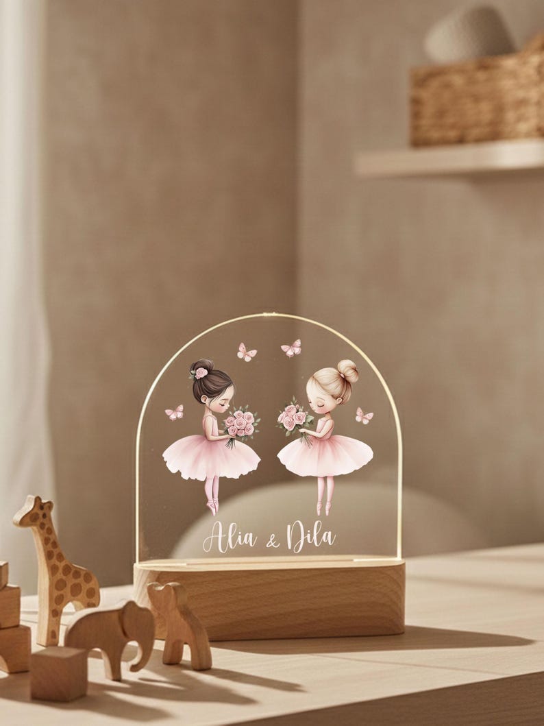 May include: A decorative night light featuring two cartoon ballerinas in pink tutus, holding bouquets, and surrounded by butterflies. The names "Alia & Dila" are written below the ballerinas. The light sits on a wooden base.