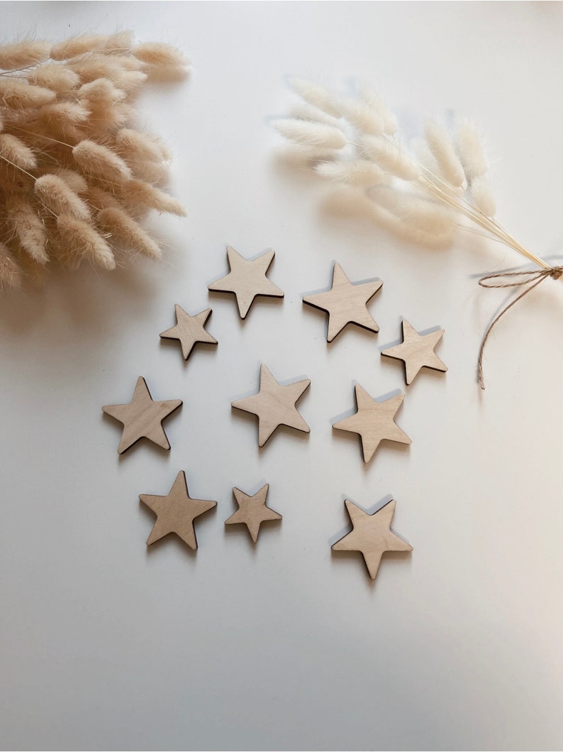 May include: A collection of ten wooden stars in various sizes, arranged in a circle on a white background. The stars are a light brown color and have a natural wood finish.