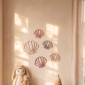 Op de afbeelding: Vijf wanddecoraties in de vorm van zeeschelpen in verschillende maten en gedempte kleuren, waaronder oudroze, mauve en lichtbruin. De schelpvormige uitsparingen hebben een waaiervormig ribbelpatroon en zijn gegroepeerd op een lichte muur.