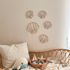 Puede incluir: Cinco decoraciones de pared de madera de color claro con forma de concha marina en varios tamaños dispuestas en una pared. Las conchas presentan un diseño recortado con líneas radiantes. El texto "by FoxForKids" está impreso verticalmente abajo a la derecha.