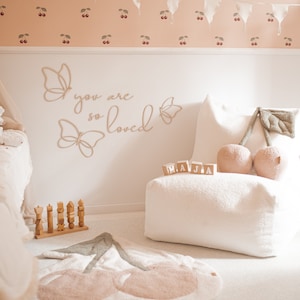 May include: A child's room with a white beanbag chair, a cherry-shaped rug, and wooden butterfly wall art. The wall features the text "you are so loved". A garland of white flags hangs above cherry-patterned wallpaper. Wooden blocks spell out "M.A.J.A."