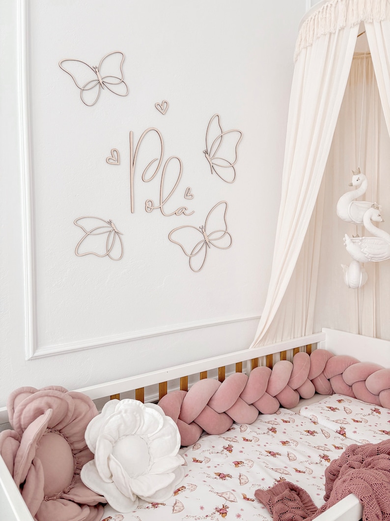 May include: A nursery scene featuring a white crib with a pink braided bumper and floral bedding. Wooden butterfly and heart wall decor spells out the name "Pola". A canopy and swan mobile add to the soft, feminine aesthetic.