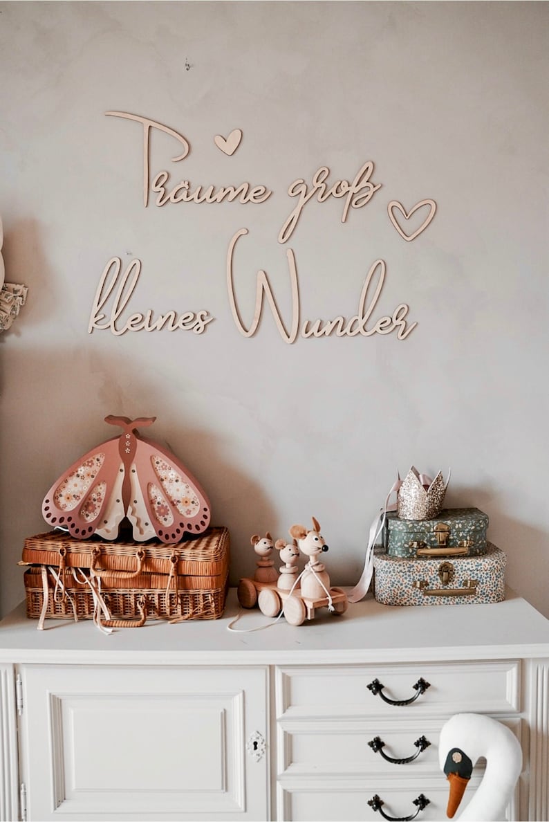 May include: Wooden wall decor with the text "Traume gro&szlig; kleines Wunder" in cursive script with two small hearts. The decor is displayed on a white wall above a white dresser with a wooden butterfly nightlight, a wooden toy train, and a white swan figurine.