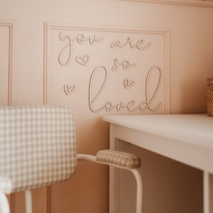 May include: A white desk with a light pink wall behind it. The wall has a gold polka dot pattern. The words "you are so loved" are written on the wall in cursive with hearts. A white chair with a gray and white checkered pattern is in front of the desk.