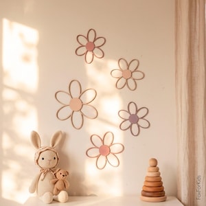 Op de afbeelding: Vijf wanddecoraties in de vorm van bloemen met dunne, afgeronde bloemblaadjes en massieve ronde centra in tinten oudroze, mauve en beige. De bloemen zijn in een groep op een effen muur gerangschikt en hebben een eenvoudig, open ontwerp.