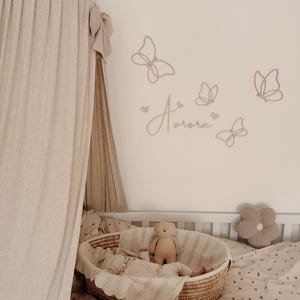 May include: A nursery scene with a white crib, a woven basket bassinet, and a beige canopy. The wall features butterfly cutouts and the name "Aurora". A teddy bear and a flower-shaped pillow are also visible.