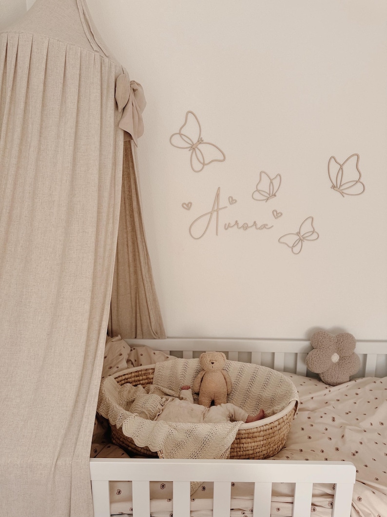 May include: A nursery scene with a white crib, a woven basket bassinet, and a beige canopy. The wall features butterfly cutouts and the name "Aurora". A teddy bear and a flower-shaped pillow are also visible.