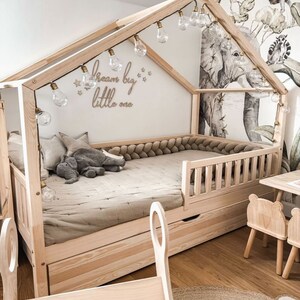 K&ouml;nnte beinhalten: Ein h&ouml;lzerner Bettrahmen in Form eines Hauses mit einer Lichterkette, die dar&uuml;ber h&auml;ngt. Das Bett hat eine hellbraune Matratze und einen grauen Stoffelefanten darauf. Die Wand hinter dem Bett hat einen wei&szlig;en Hintergrund mit den Worten "Dream big little one" in Braun geschrieben. Die Wand hat auch eine Tapete mit Dschungel-Motiv.