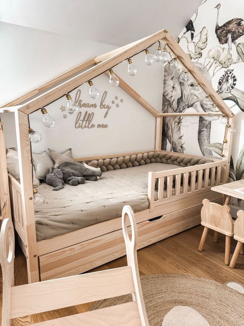 K&ouml;nnte beinhalten: Ein h&ouml;lzerner Bettrahmen in Form eines Hauses mit einer Lichterkette, die dar&uuml;ber h&auml;ngt. Das Bett hat eine hellbraune Matratze und einen grauen Stoffelefanten darauf. Die Wand hinter dem Bett hat einen wei&szlig;en Hintergrund mit den Worten "Dream big little one" in Braun geschrieben. Die Wand hat auch eine Tapete mit Dschungel-Motiv.