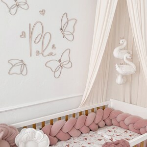 May include: A nursery scene featuring a white crib with a pink braided bumper and a patterned sheet. Wooden butterfly wall decor and the name "Pola" are above the crib. A canopy and swan mobile add to the room's soft aesthetic.
