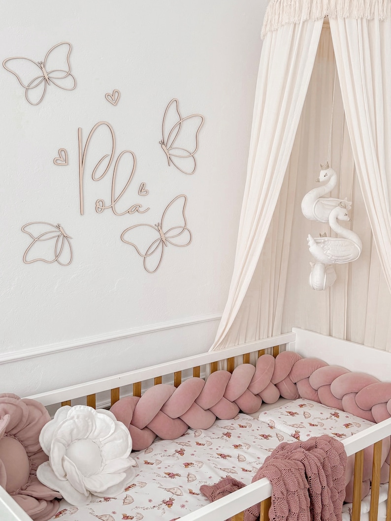 May include: A nursery scene featuring a white crib with a pink braided bumper and a patterned sheet. Wooden butterfly wall decor and the name "Pola" are above the crib. A canopy and swan mobile add to the room's soft aesthetic.