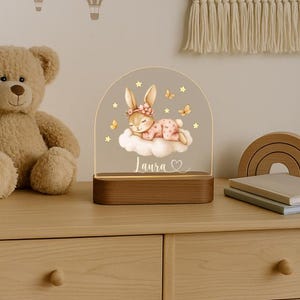 May include: A decorative night light featuring a sleeping bunny on a cloud, with the name "Laura" and a heart. The light has a wooden base and a clear acrylic top with stars and butterflies. A teddy bear and hot air balloon wall decals are in the background.