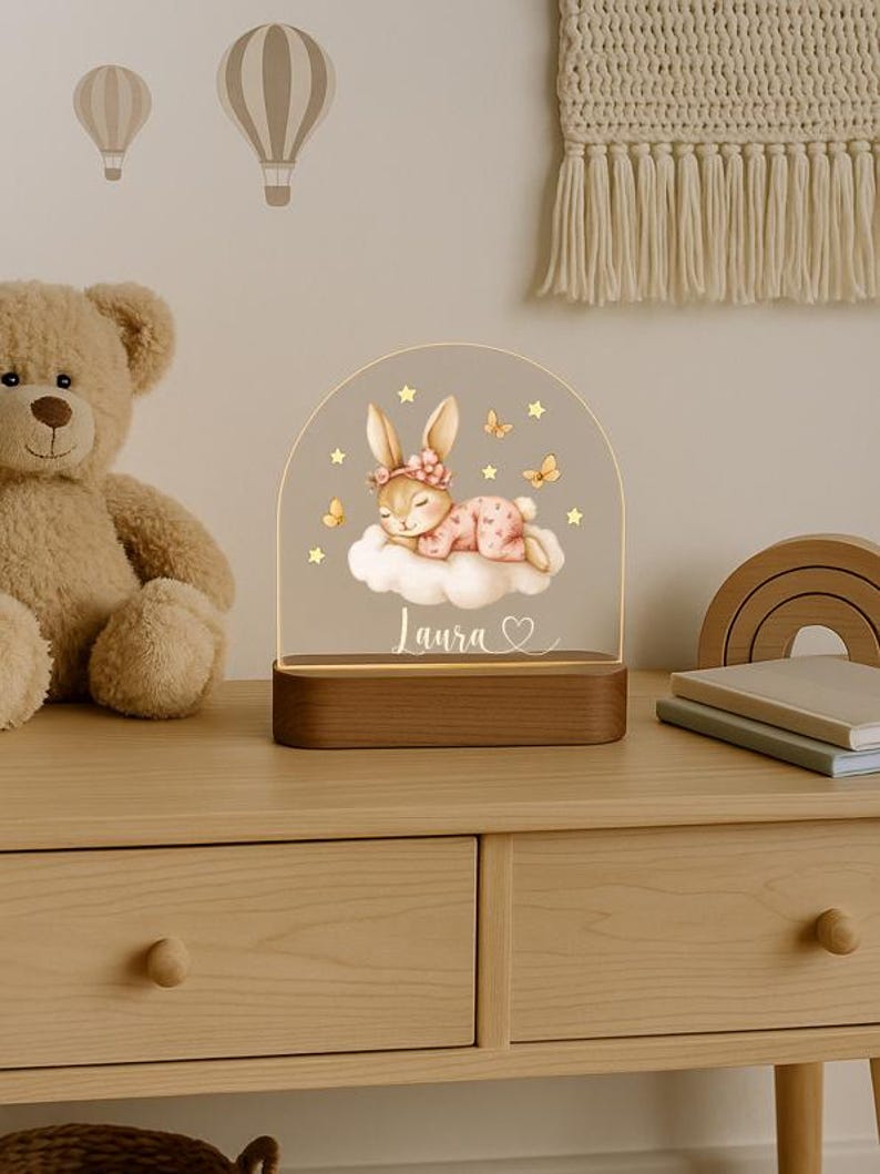 May include: A decorative night light featuring a sleeping bunny on a cloud, with the name "Laura" and a heart. The light has a wooden base and a clear acrylic top with stars and butterflies. A teddy bear and hot air balloon wall decals are in the background.