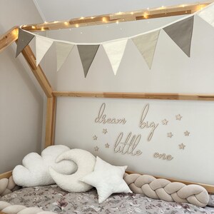 K&ouml;nnte beinhalten: Ein Holzbett mit einem wei&szlig;en und beigefarbenen Stoffbaldachin und einer Lichterkette. Das Bett ist mit einem geflochtenen, beigen Sto&szlig;f&auml;nger, wei&szlig;en Wolken- und Halbmondkissen und einem wei&szlig;en Sternkissen dekoriert. Die Wand hinter dem Bett hat ein Holzschild mit der Aufschrift "dream big little one" mit kleinen Holzsternen.