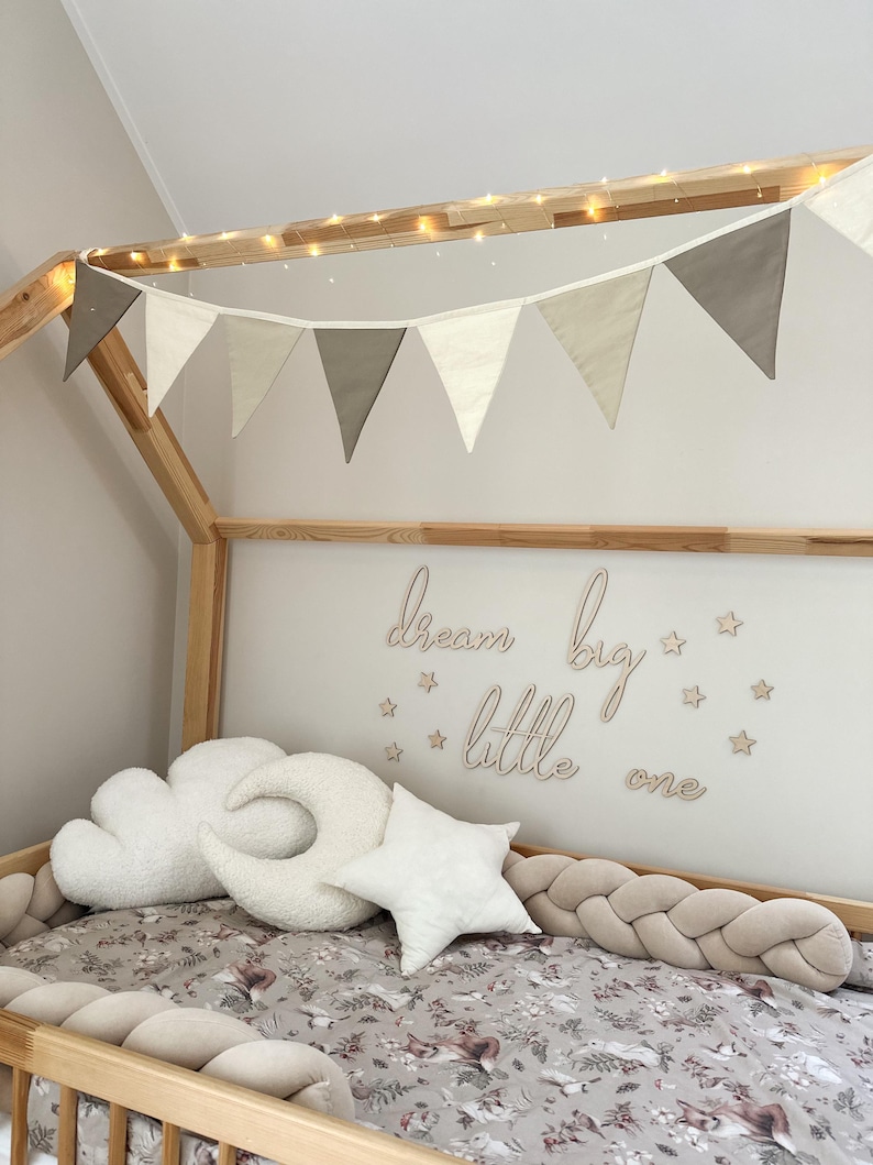 K&ouml;nnte beinhalten: Ein Holzbett mit einem wei&szlig;en und beigefarbenen Stoffbaldachin und einer Lichterkette. Das Bett ist mit einem geflochtenen, beigen Sto&szlig;f&auml;nger, wei&szlig;en Wolken- und Halbmondkissen und einem wei&szlig;en Sternkissen dekoriert. Die Wand hinter dem Bett hat ein Holzschild mit der Aufschrift "dream big little one" mit kleinen Holzsternen.