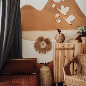 May include: A cozy nursery with a warm color palette. Features a wooden bed with a rust-colored quilt, a woven pear-shaped basket, and a wooden cabinet. The wall has a mural of a mountain scene with wooden geese.