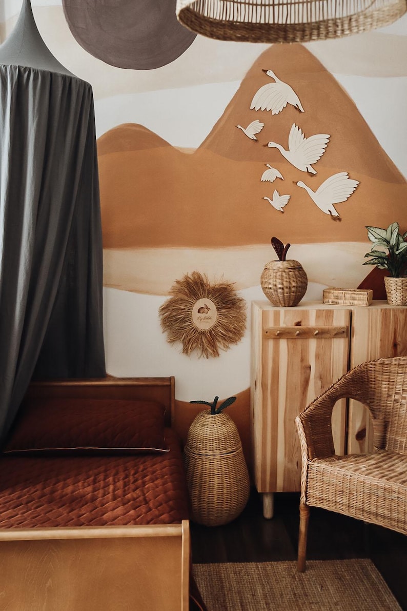 May include: A cozy nursery with a warm color palette. Features a wooden bed with a rust-colored quilt, a woven pear-shaped basket, and a wooden cabinet. The wall has a mural of a mountain scene with wooden geese.