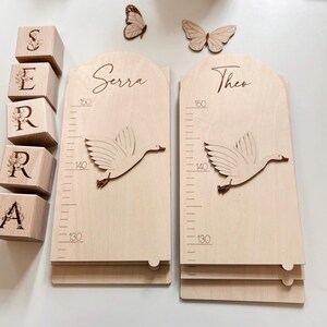 K&ouml;nnte beinhalten: Wachstumstafel aus Holz mit einem fliegenden G&auml;nsemotiv, misst bis zu 150 cm. Die Tafel ist mit den Namen "Serra" und "Theo" personalisiert. Holzbl&ouml;cke buchstabieren den Namen "SERRA". Dekorative Schmetterlinge sind ebenfalls vorhanden.